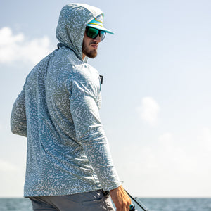 Man wearing a light gray hoodie and sunglasses, standing on a boat with water and sky in the background.
