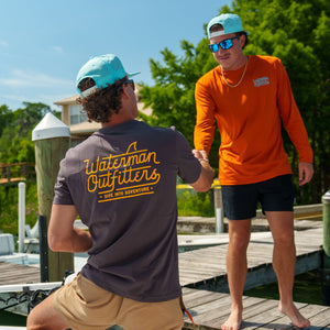 Two men on a dock, one wearing a 'Waterman Outfitters' shirt, with a boat and greenery in the background.
