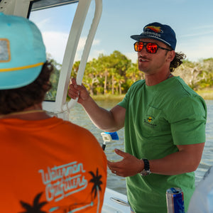 Two men on a boat, one wearing a green shirt and sunglasses, the other in an orange shirt with palm tree design.