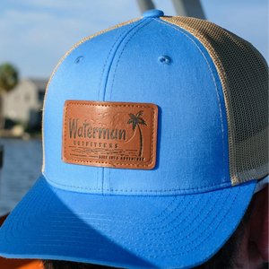 Blue cap with a leather patch labeled 'Waterman Outfitters' worn by a person outdoors.