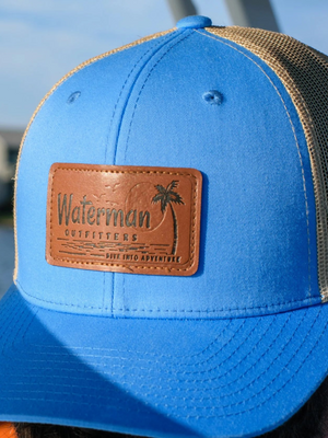 Blue cap with a leather patch labeled 'Waterman Outfitters' worn by a person outdoors.