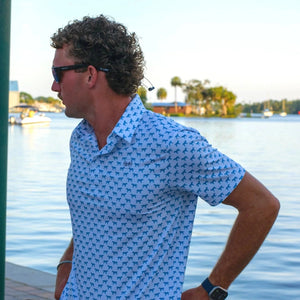 Man wearing a blue patterned shirt by a waterfront with boats and trees in the background.