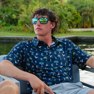 Man wearing sunglasses and a patterned shirt sitting by a body of water with greenery in the background