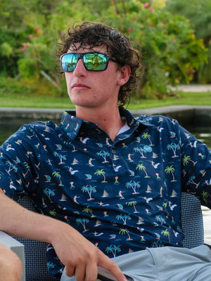 Man wearing sunglasses and a patterned shirt sitting by a body of water with greenery in the background